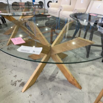 EX STAGING - 1500MM ROUND GLASS TABLE, WITH TIMBER CROSS LEG BASE, CHIPPED EDGE ON GLASS
